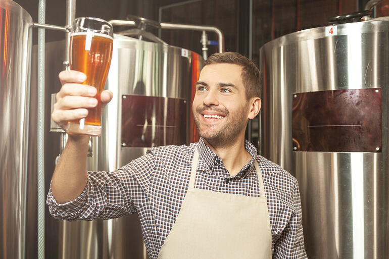 A smiling man wearing an apron holding a glass of beer at a beer plant.