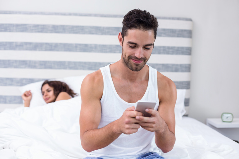 A boyfriend secretly texts on his phone while his girlfriend lies in bed, unaware of his actions