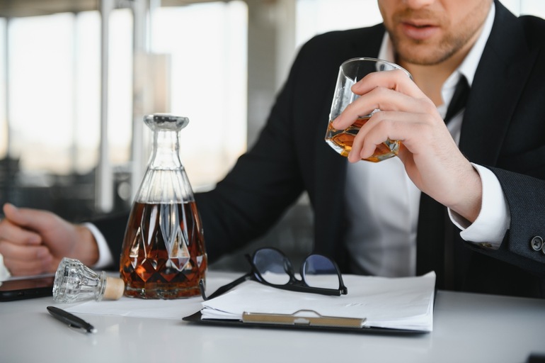 A businessman is drinking heavily at his desk with documents and a decanter nearby